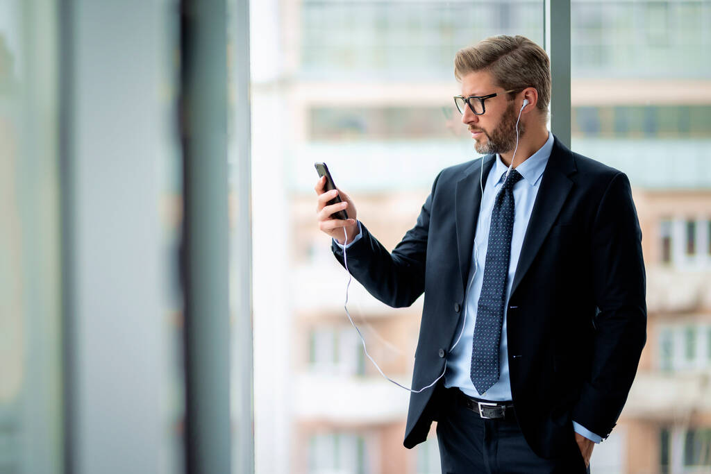 Vážný obchodník používající smartphone a sluchátka, když stojí v kanceláři. Profesionální muž s video hovorem.   - Fotografie, Obrázek