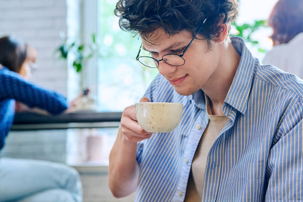 Przystojny młody kręcone włosy facet pijący filiżankę kawy w kawiarni. Młody student w okularach delektujący się cappuccino. Kultura kawy, styl życia, młodzież, koncepcja ludzi - Zdjęcie, obraz