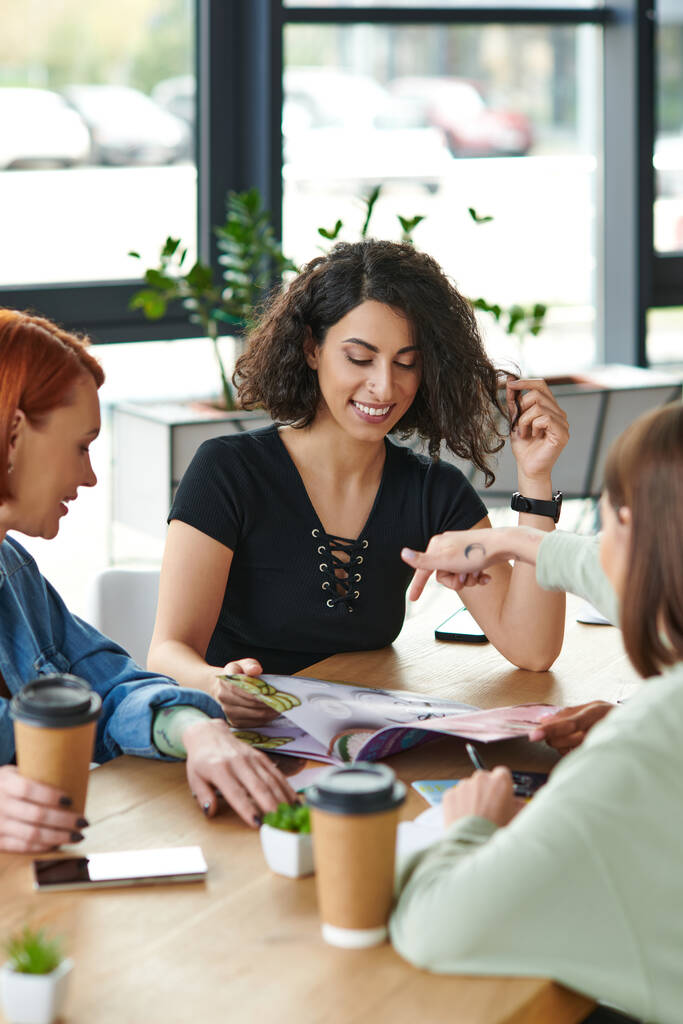 young woman pointing at magazine near smiling multicultural girlfriends and paper cups with coffee to go on table in women club, common interests and knowledge-sharing concept - Photo, Image