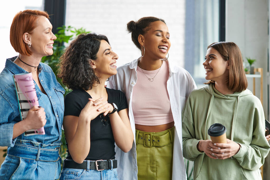 junge und glückliche Frau mit Coffee to go beim Anblick stilvoller multiethnischer Freundinnen, die im Fraueninteressenclub stehen und lachen, gegenseitige Unterstützung, Solidarität und Vertrauenskonzept - Foto, Bild