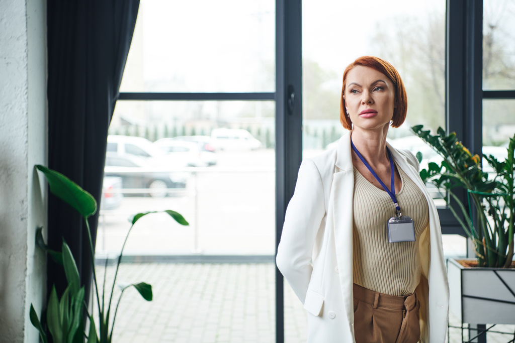 thoughtful psychologist with red hair and name badge standing in white blazer, thinking and looking away in consulting room, professional help and supportive therapy - Photo, Image