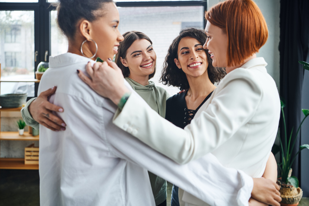 boldog és optimista multikulturális barátnők ölelkeznek vörös hajú motivációs edzővel és egymásra mosolyognak a támogató terápia alatt a konzultációs szobában, a női egység és támogatás koncepciójában - Fotó, kép