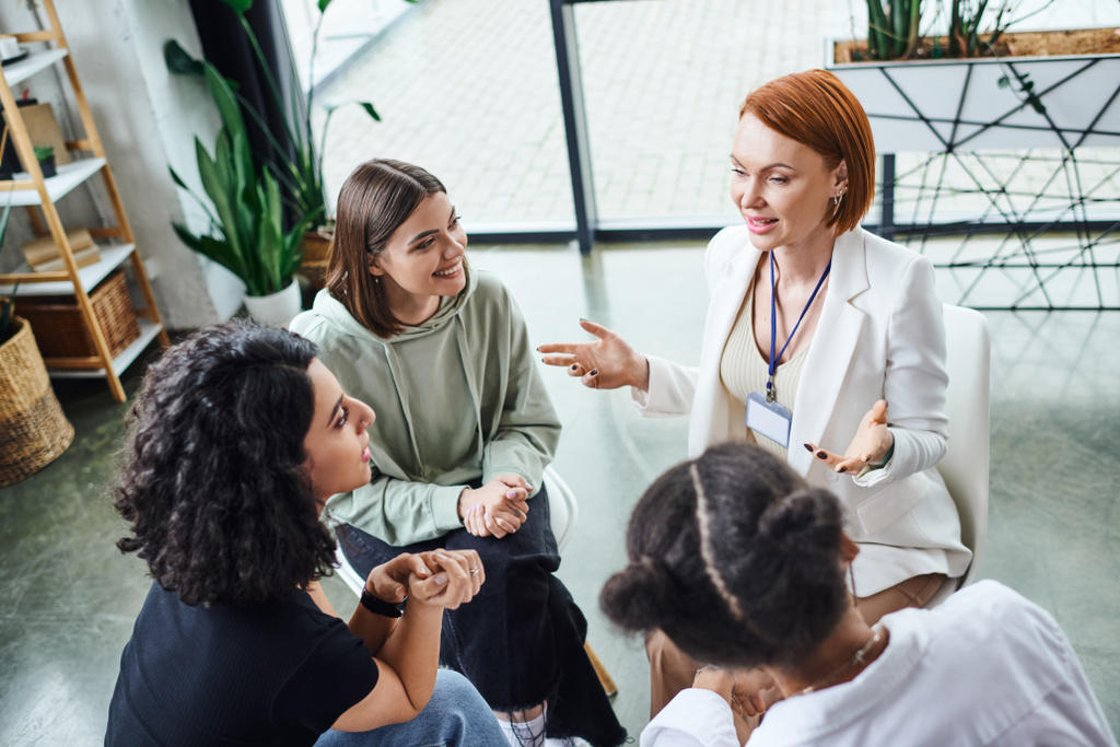 high angle view of redhead psychologist pointing with hands and talking to smiling multicultural women during motivation therapy in consulting room, friendship and mental wellness concept - Photo, Image