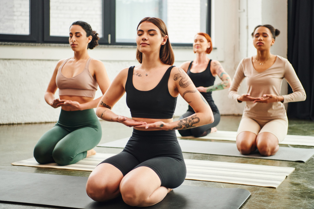wytatuowany trener jogi medytujący zamkniętymi oczami siedząc w pozycji grzmotu w pobliżu zróżnicowanej grupy wieloetnicznych kobiet, wewnętrznej koncepcji spokoju i świadomości ciała - Zdjęcie, obraz