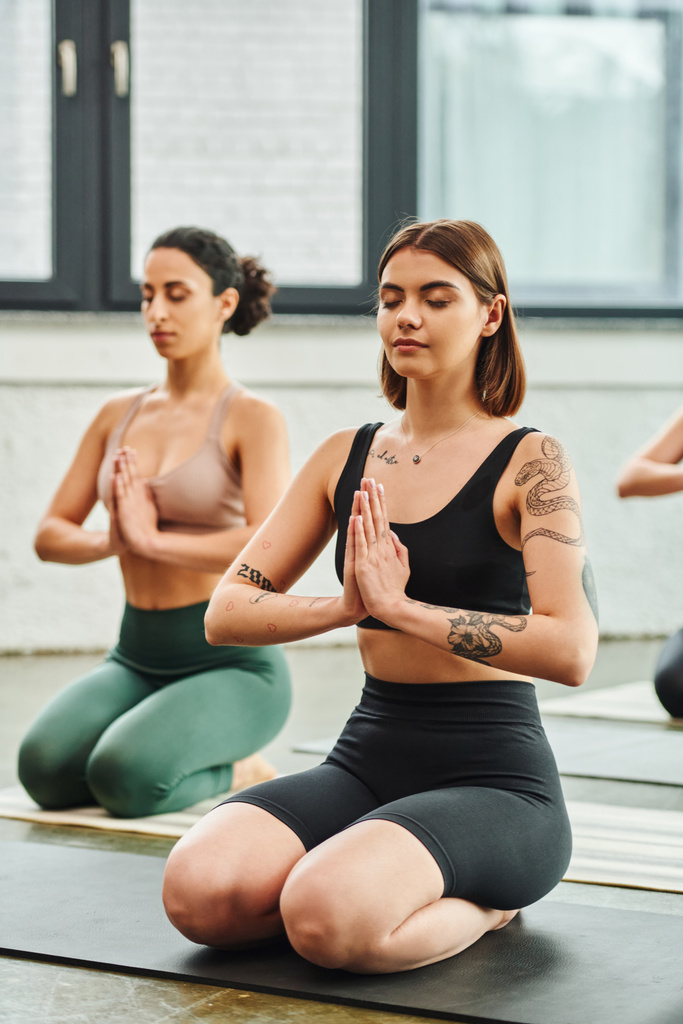 young tattooed woman sitting in thunderbolt pose and meditating with closed eyes and praying hands near multiracial girlfriend on blurred background, inner peace and body awareness concept - Photo, Image