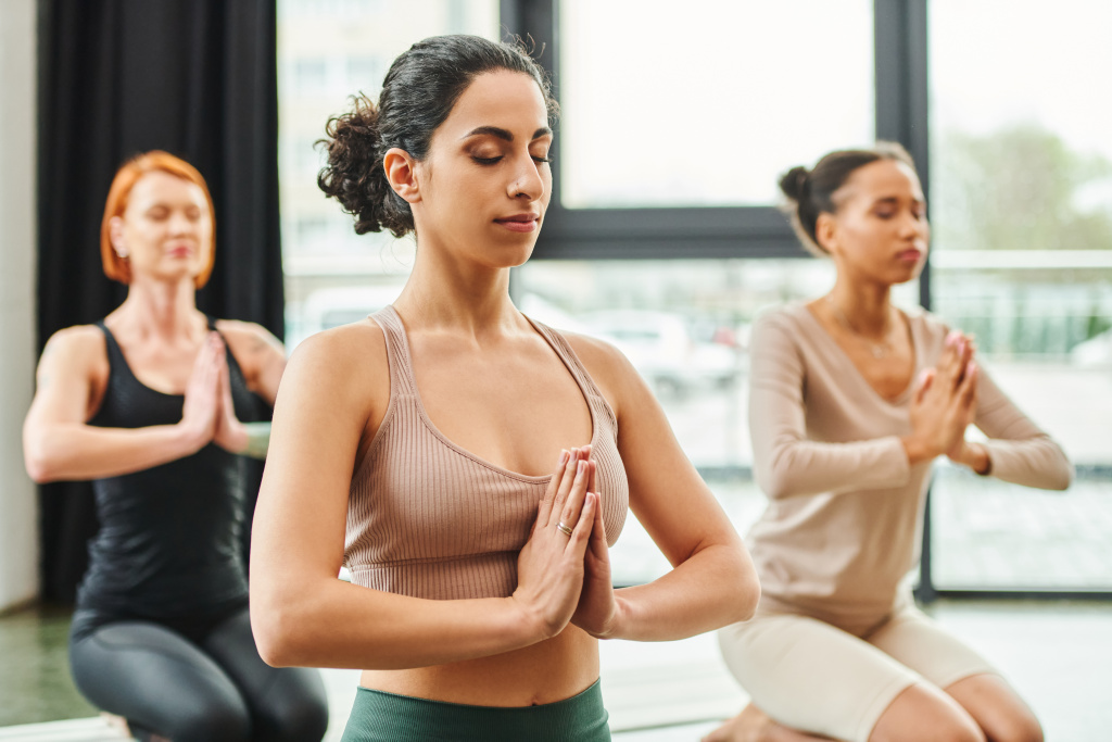 multiethnische Frau in Sportkleidung meditiert mit geschlossenen Augen und betenden Händen während eines Yoga-Kurses in der Nähe multiethnischer Freundinnen auf verschwommenem Hintergrund, innerem Frieden und Körperwahrnehmungskonzept - Foto, Bild