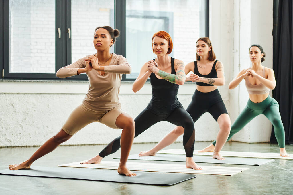 full length of multiethnic female friends in sportswear standing in crescent lunge pose with clenched hands while meditating during yoga class, harmony and mental health concept - Photo, Image
