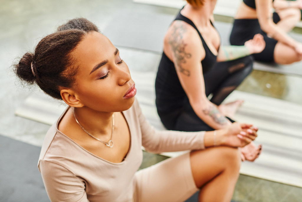 hoge hoek uitzicht op jonge Afrikaans-Amerikaanse vrouw met gesloten ogen mediteren tijdens yogales naast vrouwelijke vrienden zitten in gemakkelijke pose op wazig achtergrond, harmonie en wellness concept - Foto, afbeelding