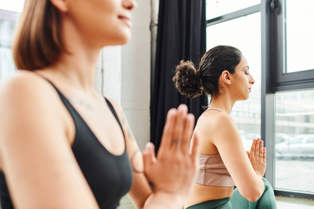 donna multirazziale seduta con gli occhi chiusi e meditare con le mani in preghiera durante la lezione di yoga vicino fidanzata in primo piano offuscata, armonia e concetto di benessere - Foto, immagini