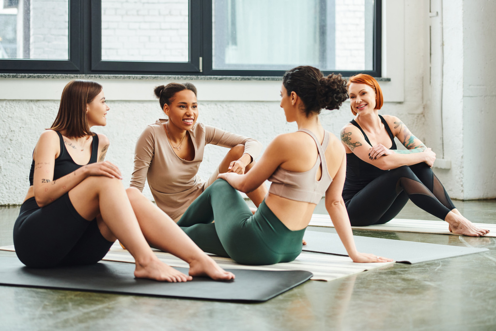 Çeşitli mutlu ve gülümseyen kadın arkadaş grupları spor kıyafetleri içinde yoga minderleri üzerinde oturup spor salonu, arkadaşlık, ahenk ve zihinsel sağlık konseptiyle konuşuyorlar. - Fotoğraf, Görsel