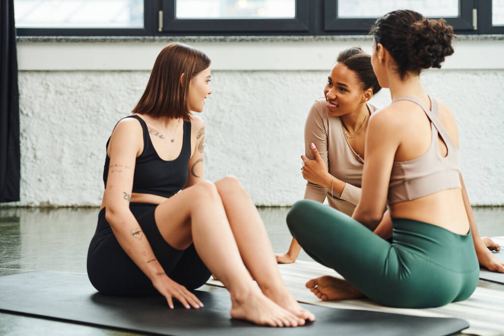 jong en positief Afrikaans amerikaanse vrouw in sportkleding zitten op yoga mat en praten met getatoeëerde vriendin tijdens yoga les in sportschool, vriendschap, harmonie en geestelijke gezondheid concept - Foto, afbeelding