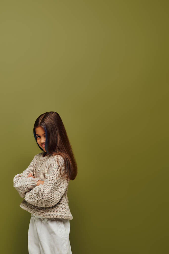 Offended and stylish preteen girl with colored hair wearing knitted sweater and crossing arms while standing and posing isolated on green, fashion-forward preteen with sense of style - Photo, Image