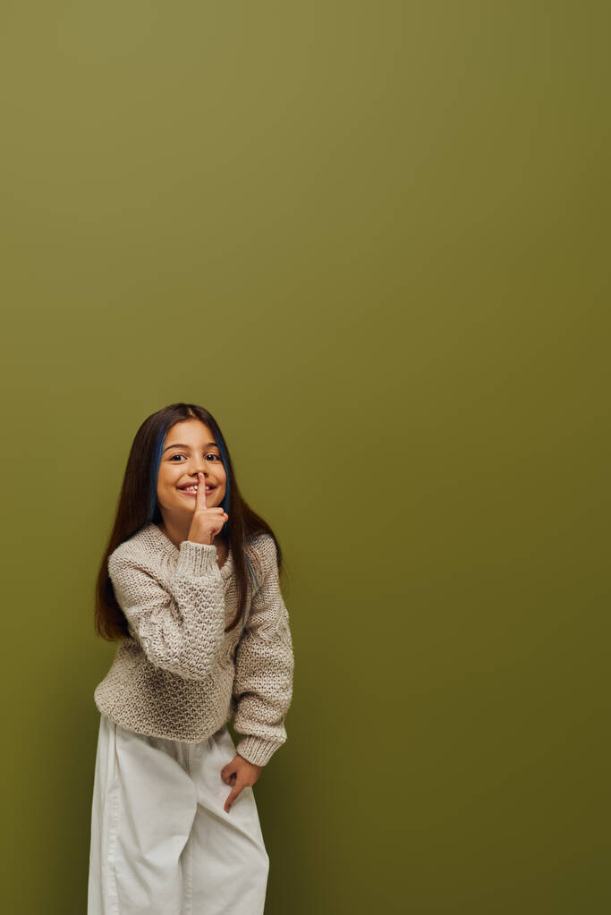 Cheerful and stylish preteen girl with dyed hair in knitted sweater showing secret gesture and looking at camera while standing on green background, modern and hip preteen fashion - Photo, Image