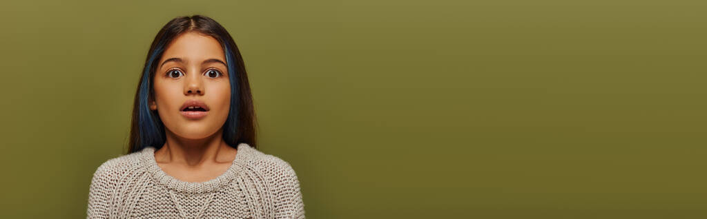 Портрет шокированного ребенка-подростка с окрашенными волосами в современный вязаный свитер и смотрящего в камеру, стоящего изолированно на зеленом, баннер с копировальным пространством, современная детская мода - Фото, изображение