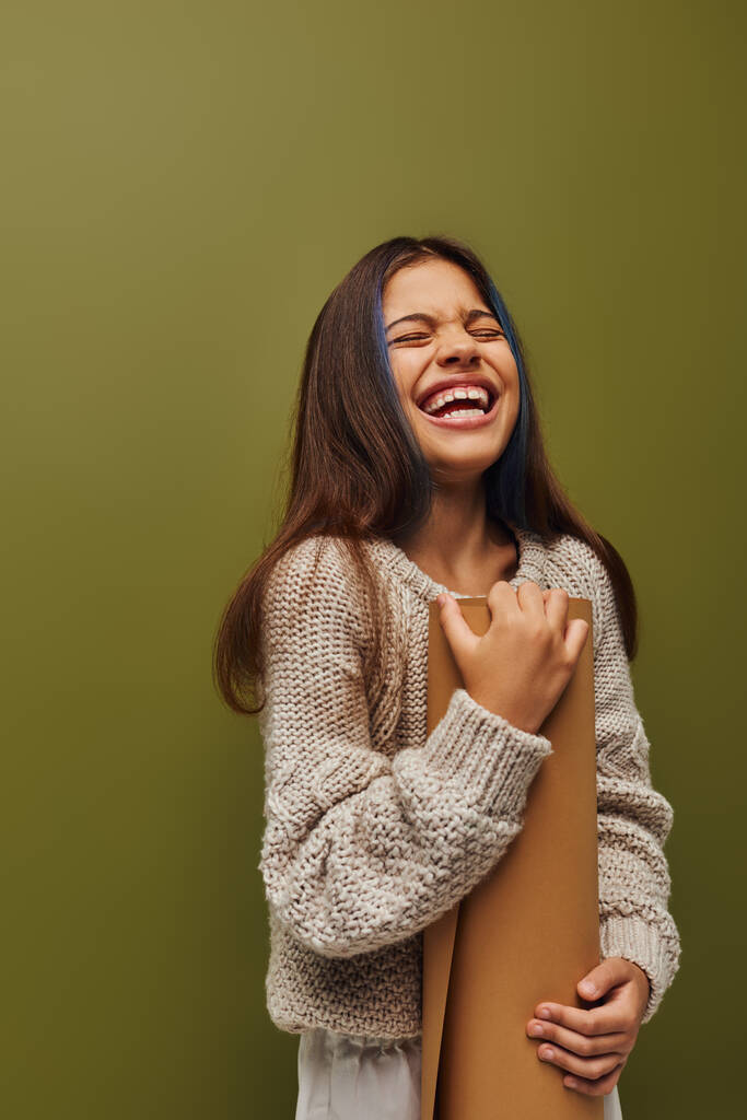Vidám és elegáns tizenéves lány modern kötött pulóver és őszi ruhát tartó hengerelt papír és nevetés közben elszigetelt zöld, lány sugárzó őszi vibrálás koncepció - Fotó, kép