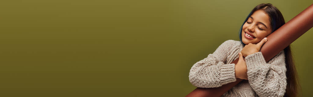 Vrolijk en stijlvol preadolescent meisje met geverfd haar draagt gebreide trui terwijl knuffelen opgerold papier en staan geïsoleerd op groen, meisje stralen herfst vibes, banner met kopieerruimte - Foto, afbeelding