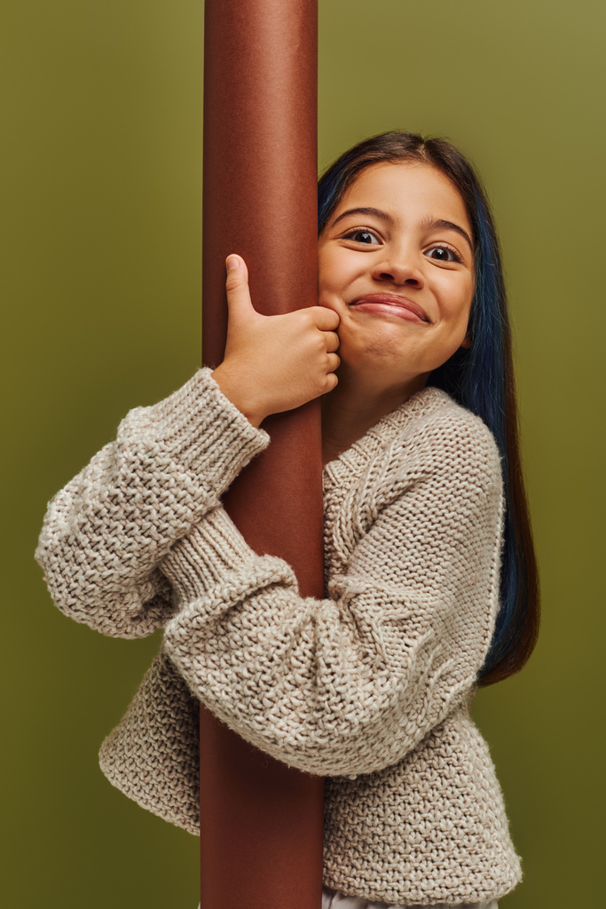 Retrato de animado e sorridente menina pré-adolescente na moda em suéter de malha abraçando papel laminado e olhando para a câmera enquanto estava isolado em verde, menina irradiando conceito vibrações de outono - Foto, Imagem