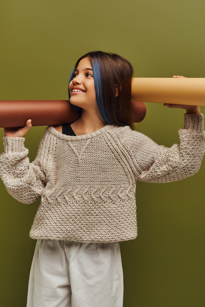 Позитивна дівчина-підліток з фарбованим волоссям в модному осінньому вбранні і тримає рулонні папери, дивлячись ізольовано на зеленій, осінній моді концепції - Фото, зображення