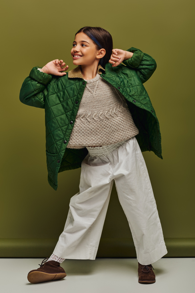 Longitud completa de niño preadolescente sonriente y de moda en traje de otoño y suéter de punto mirando hacia otro lado mientras posa y de pie sobre un fondo verde, moda infantil moderna para el concepto de preadolescentes - Foto, imagen