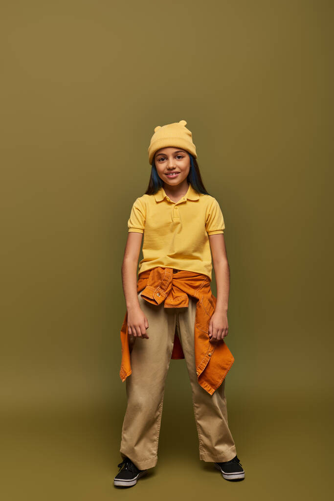 Volledige lengte van lachende en stijlvolle preadolescent meisje met geverfd haar dragen stedelijke outfit en gele hoed terwijl staan op kaki achtergrond, stijlvol meisje in moderne outfit concept - Foto, afbeelding