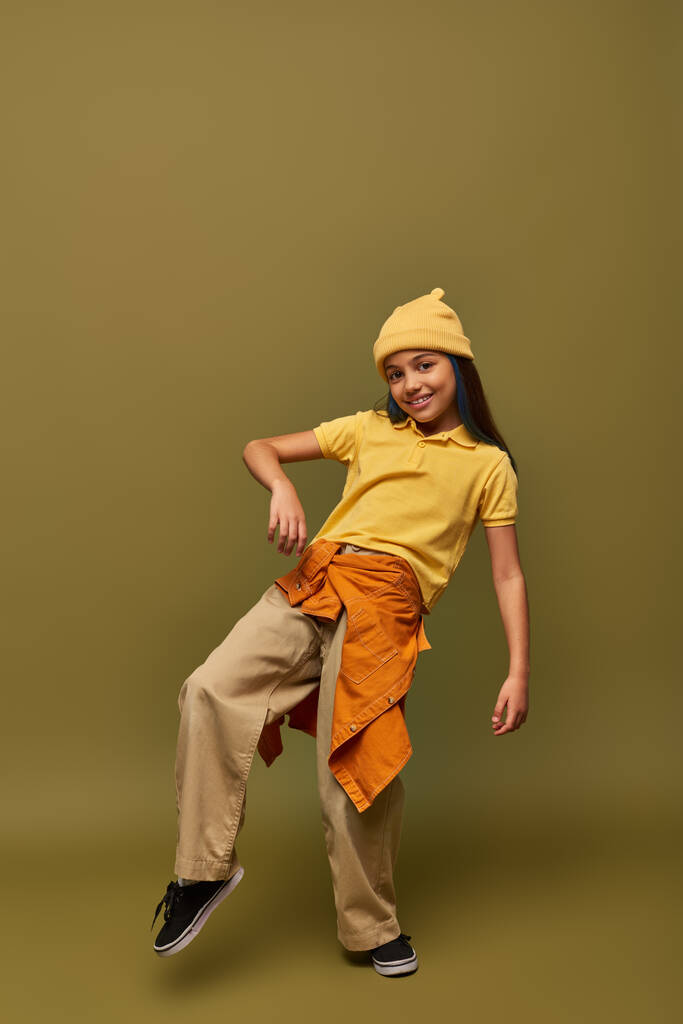 Full length of trendy and cheerful preadolescent girl in urban outfit and yellow hat posing while looking at camera on khaki background, stylish girl in modern outfit concept - Photo, Image
