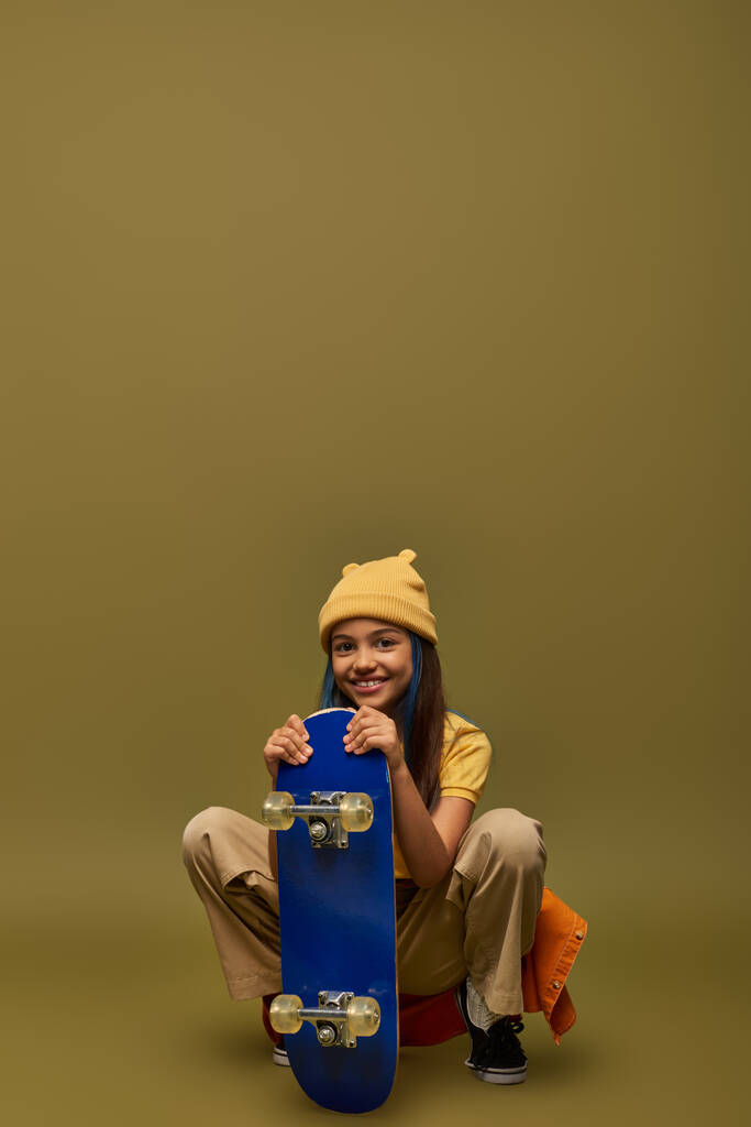 Menina pré-adolescente elegante com cabelo tingido usando chapéu amarelo e roupa urbana enquanto posando com skate e sentado em fundo cáqui, menina elegante no conceito de roupa moderna - Foto, Imagem