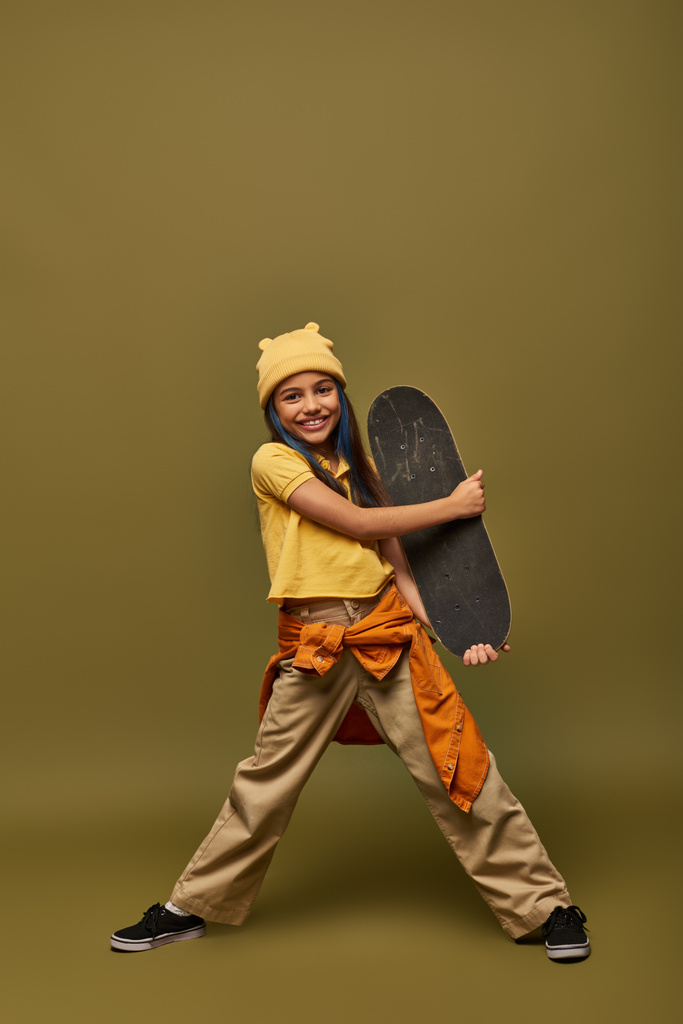 Piena lunghezza di positivo preadolescente ragazza in cappello giallo e vestito urbano in possesso di skateboard e guardando la fotocamera su sfondo kaki, ragazza alla moda nel concetto di abbigliamento moderno - Foto, immagini