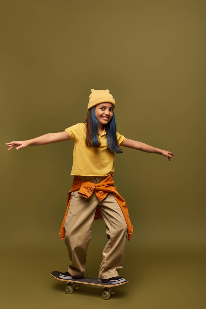 Радостная девочка-подросток с окрашенными волосами в стильный городской наряд и шляпу, стоя на скейтборде и глядя на камеру на фоне хаки, девушка с прохладным уличным видом - Фото, изображение