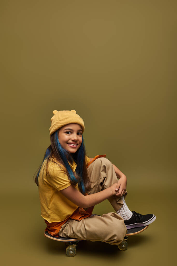 Χαμογελώντας preteen και κομψό παιδί με βαμμένα μαλλιά φορώντας αστική στολή και καπέλο κοιτάζοντας κάμερα, ενώ κάθεται σε skateboard σε χακί φόντο, κορίτσι με δροσερό στυλ δρόμο ματιά - Φωτογραφία, εικόνα