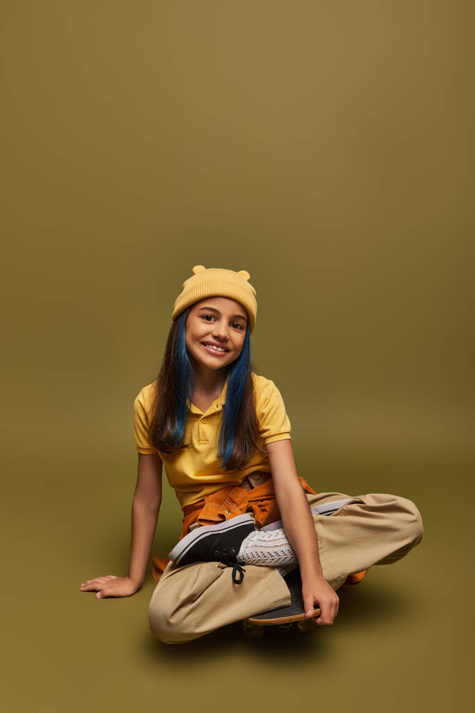 Pozitivní a módní pre-teen dívka s barvenými vlasy na sobě městské oblečení a žlutý klobouk při pohledu na kameru a sedí na skateboardu na khaki pozadí, dívka s cool street style look - Fotografie, Obrázek