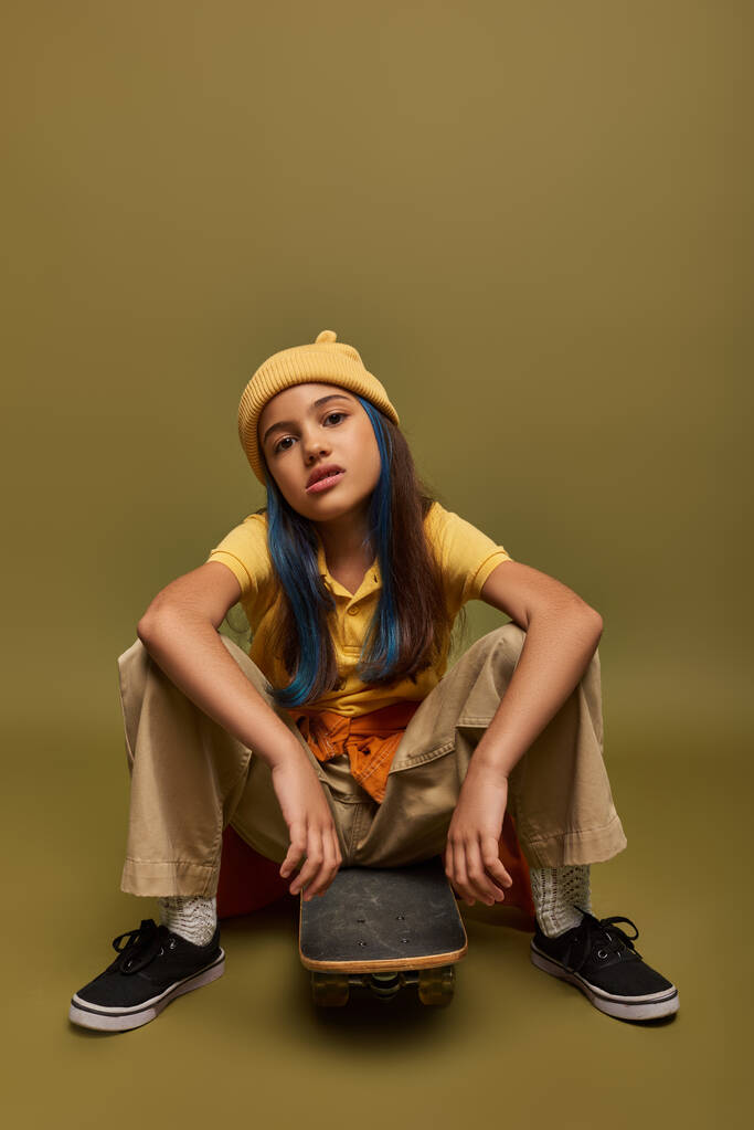Selbstbewusstes und seriöses Mädchen mit gefärbten Haaren, das urbanes Outfit und Hut trägt, auf Skateboard sitzt und auf khakifarbenem Hintergrund in die Kamera schaut, Mädchen mit coolem Street-Style-Look - Foto, Bild