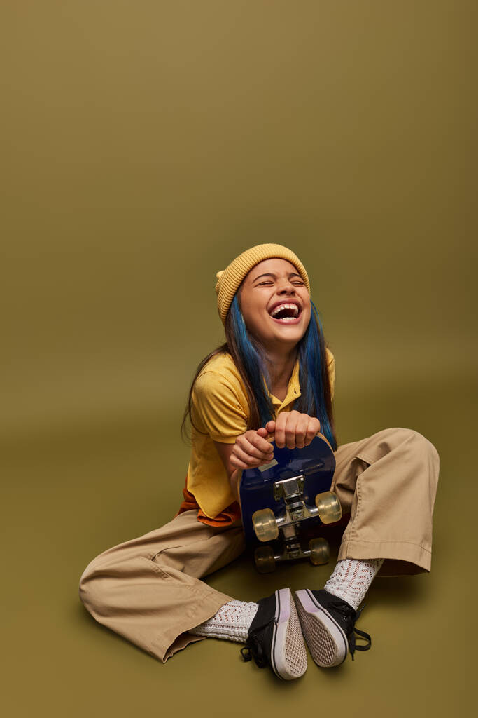 Rire et élégant fille préadolescente avec des cheveux colorés portant tenue urbaine et chapeau jaune tenant skateboard et assis sur fond kaki, fille avec look de style de rue cool - Photo, image