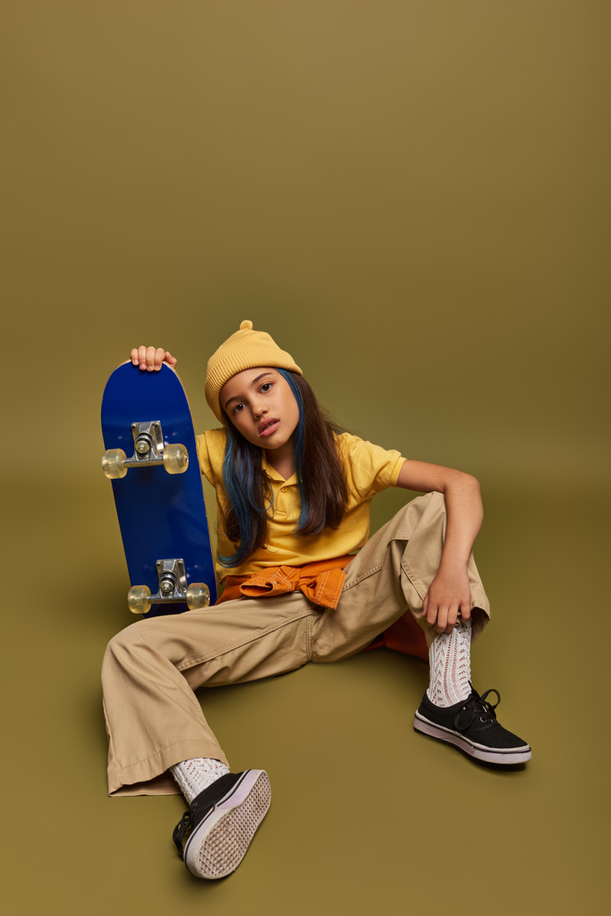 Full length of confident and fashionable preadolescent child with colored hair wearing urban clothes and hat while holding skateboard on khaki background, girl with cool street style look - 写真・画像