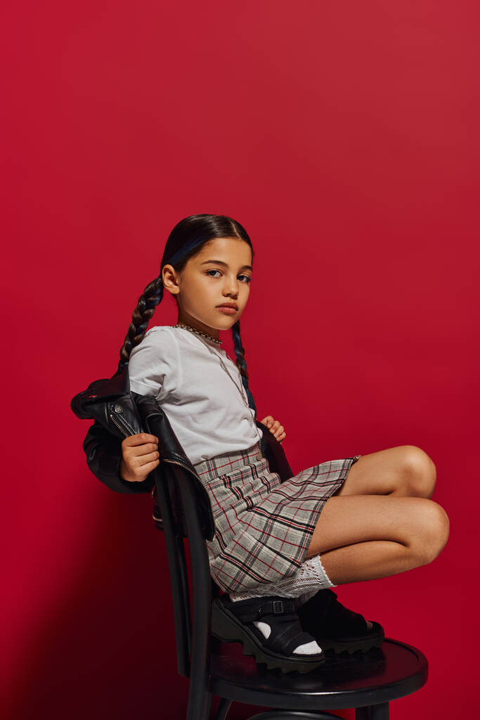 Трендовая дошкольница с прической, позирующая в кожаной куртке и чеканной юбке и смотрящая в камеру, сидя на стуле на красном фоне, стильная концепция наряда для дошкольников - Фото, изображение