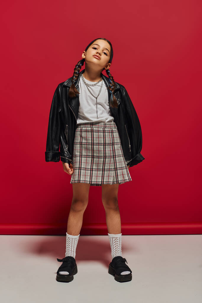 Selbstbewusstes und modisches Preteen-Mädchen mit Frisur, das Lederjacke und karierten Rock trägt, während es auf rotem Hintergrund steht und posiert, stilvolles Preteen-Outfit-Konzept - Foto, Bild