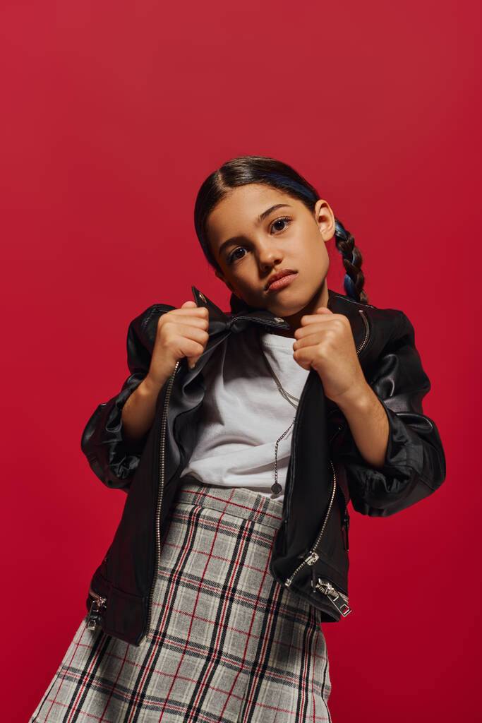 Portrait of fashionable preadolescent girl with hairstyle posing in checkered skirt while holding leather jacket and looking at camera isolated on red, stylish preteen outfit concept - Photo, Image