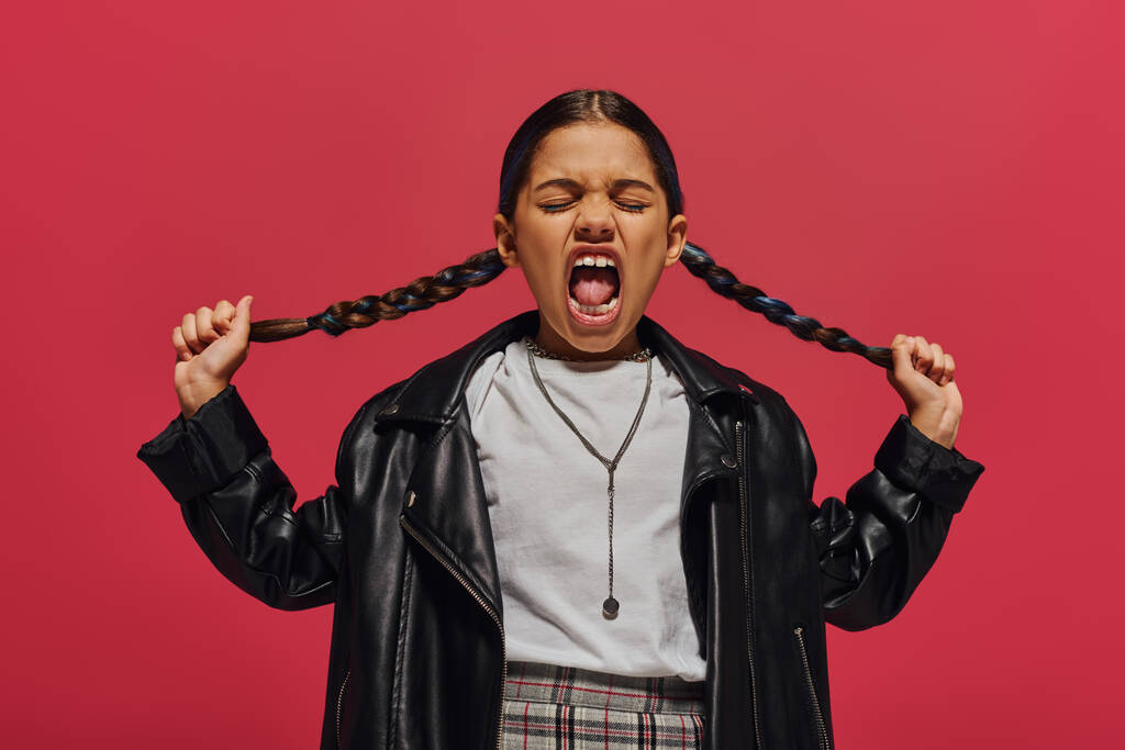 Mad and fashionable preadolescent girl screaming and touching hairstyle while posing in leather jacket and white t-shirt and standing isolated on red, girl with cool and contemporary look - 写真・画像