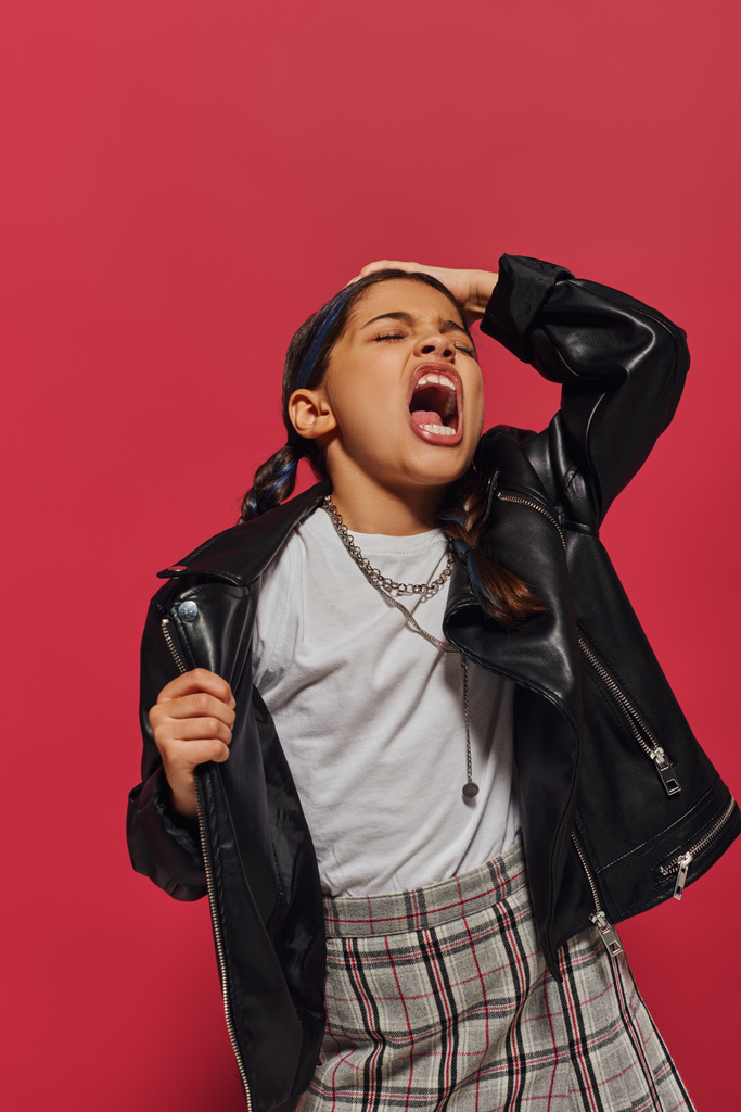 Wütend und modisch preteen Mädchen mit Frisur schreien und den Kopf berühren, während sie in Lederjacke posiert und isoliert auf rot steht, Frisur und trendiges Accessoire-Konzept - Foto, Bild