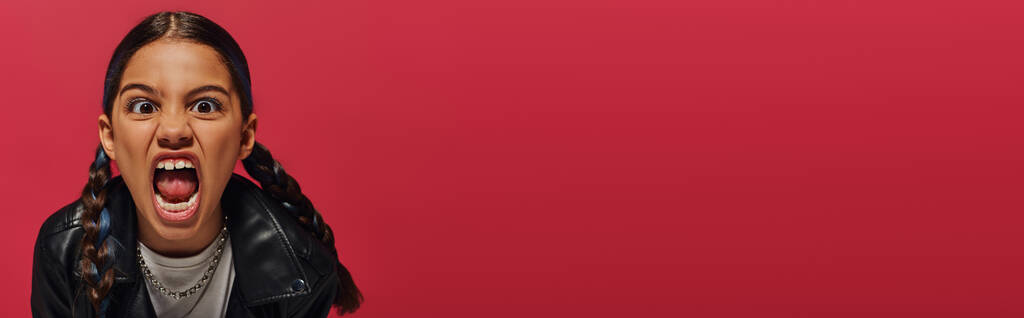 Portrait de folle et élégante fille préadolescente avec coiffure criant à la caméra et posant dans une veste en cuir isolé sur le rouge, concept de coiffure et d'accessoires à la mode, bannière  - Photo, image