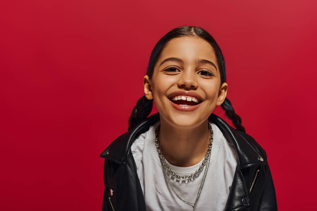 Portrait of pleased preadolescent girl with hairstyle wearing stylish leather jacket and looking at camera while posing isolated on red, hairstyle and trendy accessories concept - Photo, Image