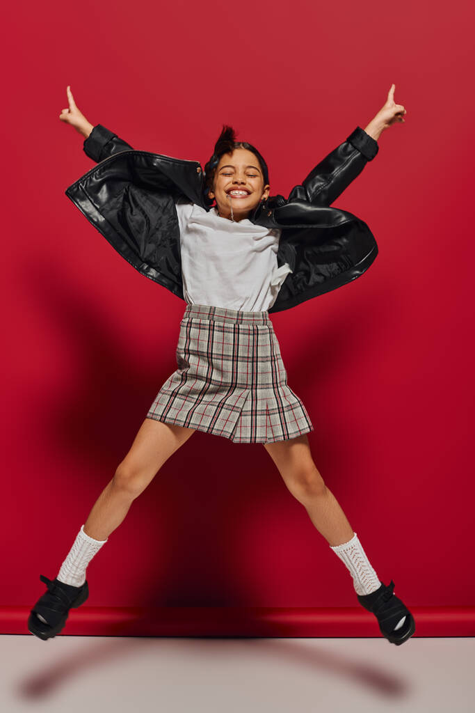 Plná délka veselý a módní dívka před dospívající v kostkované sukni a kožené bundě při skákání a ukazuje prsty na červené pozadí, účes a módní příslušenství koncepce - Fotografie, Obrázek
