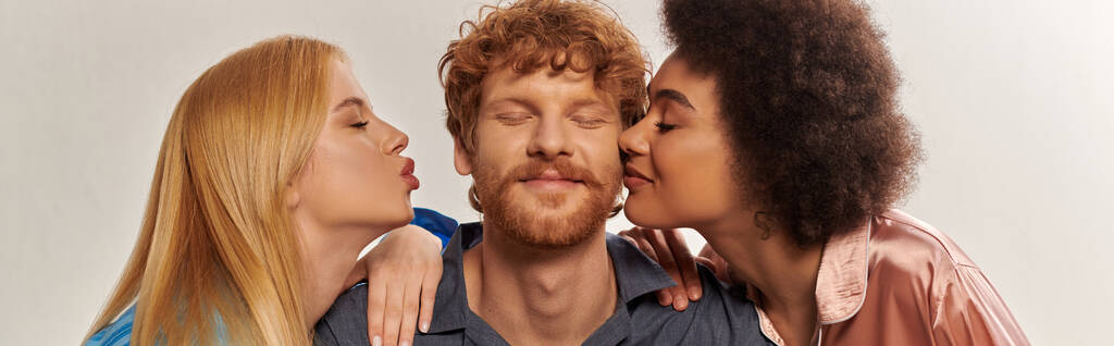 Polygamie-Konzept, offene Beziehung, Porträt von drei Erwachsenen, multikulturelle Frauen küssen glücklichen Rotschopf, polyamouröse Familie im Pyjama, kulturelle Vielfalt, Akzeptanz, Banner  - Foto, Bild