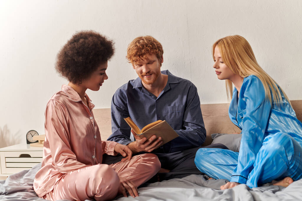 концепция открытых отношений, рыжеволосый мужчина, читающий книгу рядом со счастливыми мультикультурными женщинами в пижамах, сидящими дома на кровати, культурное разнообразие, бисексуализм, полигамия, взаимопонимание, трое взрослых  - Фото, изображение