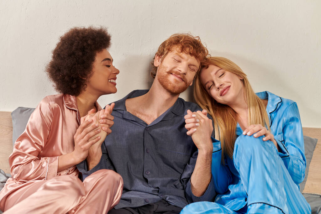 open relatie, polygamie, gelukkig drie volwassenen, roodharige man en multiculturele vrouwen in pyjama 's hand in hand in bed, culturele diversiteit, acceptatie, biseksueel, modern gezin  - Foto, afbeelding