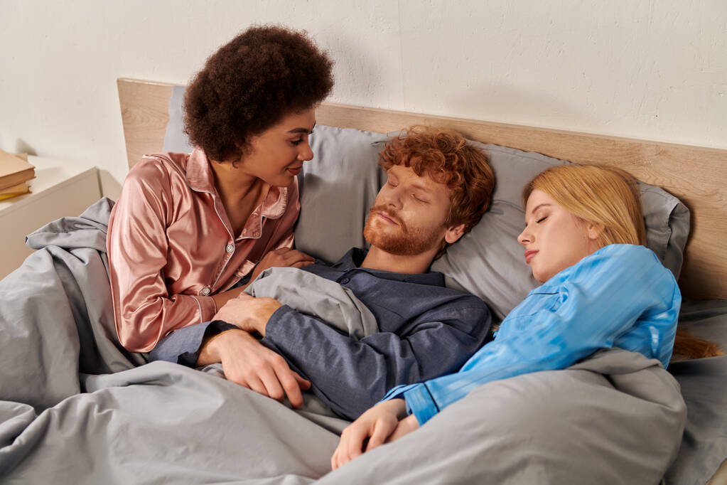 Aşk üçgeni, uyanmış Afro-Amerikalı kadın yatakta sarışın bir kadının yanındaki kızıl saçlı adama bakıyor. Çoklu aşk, geleneksel olmayan ilişkiler, çok ırklı, kültürel çeşitlilik.  - Fotoğraf, Görsel