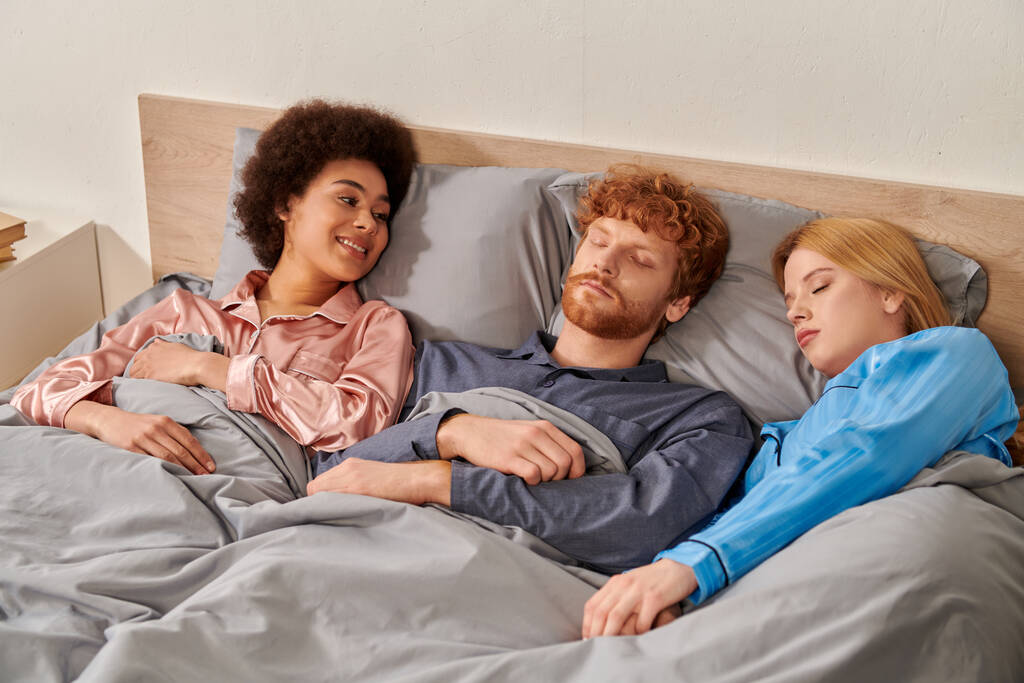 Aşk üçgeni, mutlu Afro-Amerikalı kadın. Uykulu kızıl saçlı adama bakıyor. Yatakta sarışın kadın ortağın yanında.  - Fotoğraf, Görsel