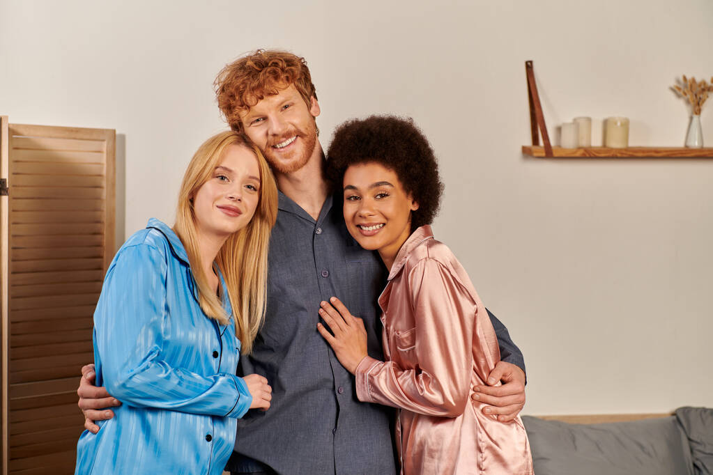 polyamory lovers in pajamas, happy man with red hair hugging interracial women at home, cultural diversity, non traditional partners, freedom in relationship, acceptance and understanding, happiness - Photo, Image