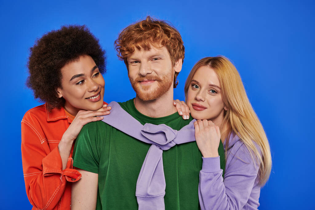 alternative relationships, polyamory, portrait of three people, young redhead man and beautiful multiracial women on blue background, studio shot, vibrant clothes, stylish attire, modern family  - Photo, Image