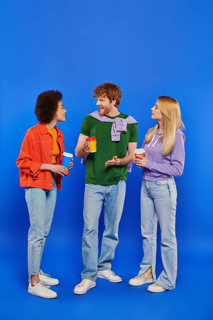polygamy people with coffee, young redhead man and beautiful multiracial women holding paper cups on blue background, studio shot, takeaway drink, polyamorous lovers, conversation  - Photo, Image