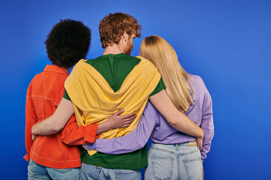 miłosny trójkąt, miłośnicy poliamii, widok z tyłu młodego rudego mężczyzny przytulającego wielorasowe kobiety na niebieskim tle, ujęcie studyjne, żywe kolory, odzież casual, stylowe stroje, nowoczesna rodzina  - Zdjęcie, obraz